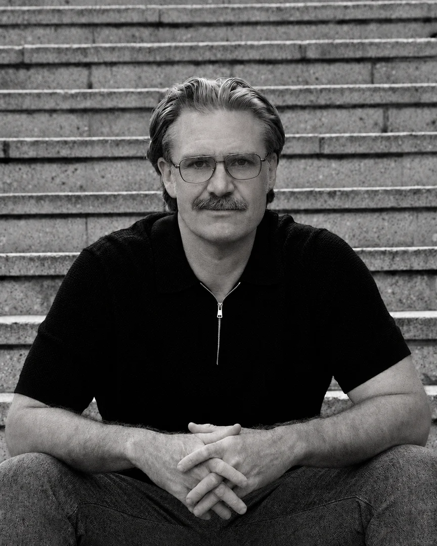 The same steps years on: seated in a black zip polo with glasses and a moustache, hands clasped between the knees; a black and white photograph.