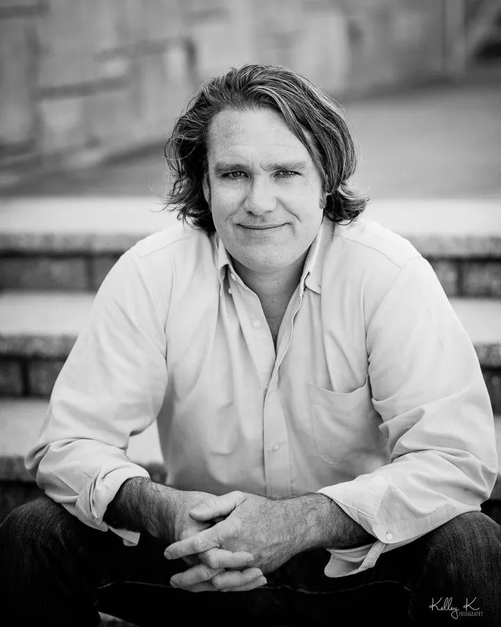 Joel Stevens, seated on stone steps in a white shirt, sleeves rolled, arms folded across the knees, smiling; a black and white photograph.